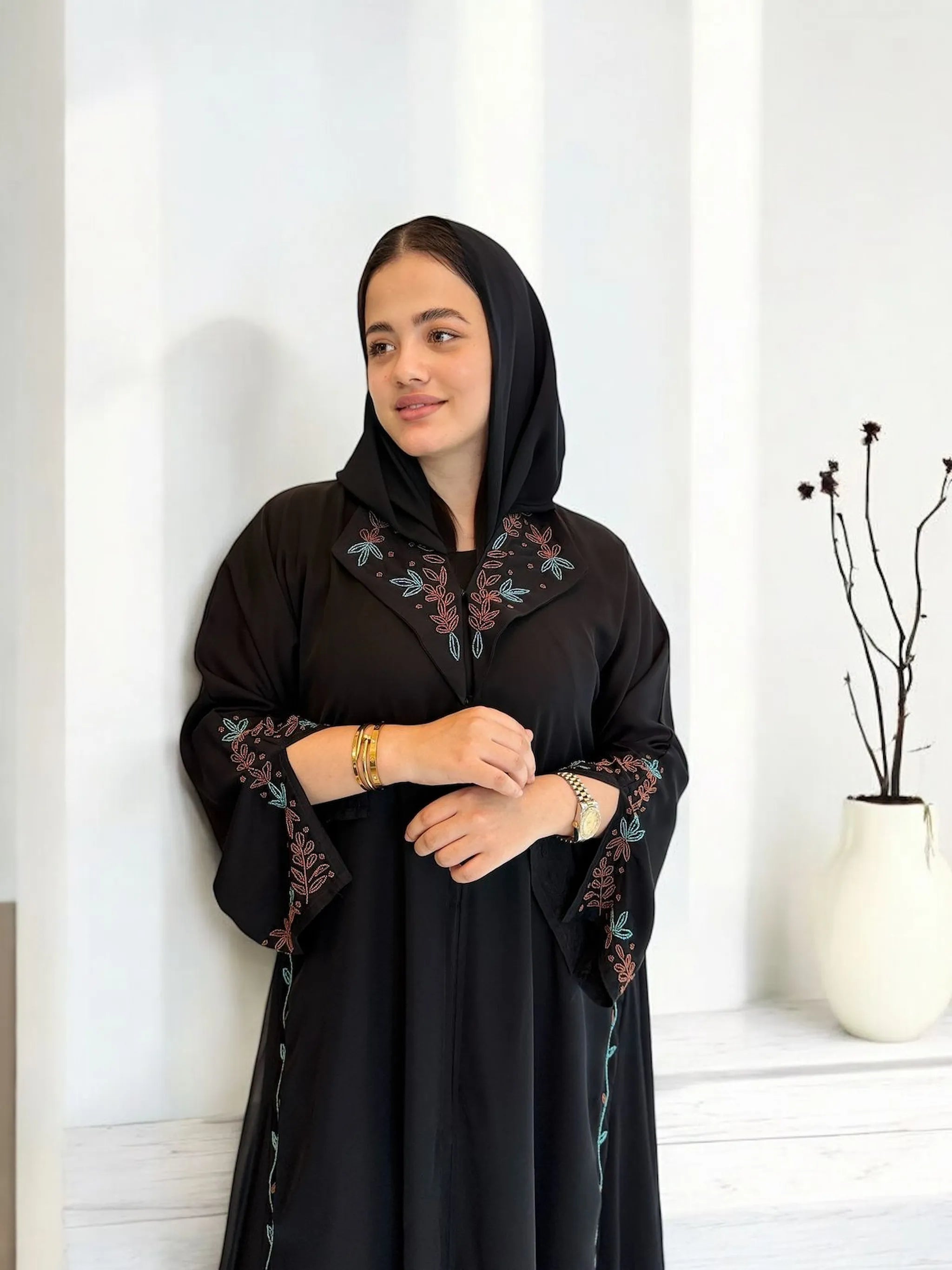 امرأة ترتدي عباية سوداء بتطريز وردي أنيق، تقف في غرفة بتصميم بسيط.
Woman wearing a black abaya with elegant pastel embroidery, standing in a minimalistic room.